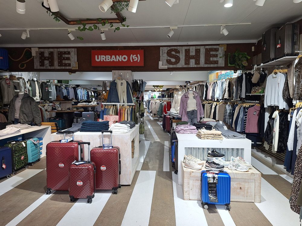 Inside a clothing store: packed rails on both sides, tables of folded clothes and trainers, mannequins and price-tagged suitcases in the foreground, under signs reading “HE”, “URBANO (5)” and “SHE”.