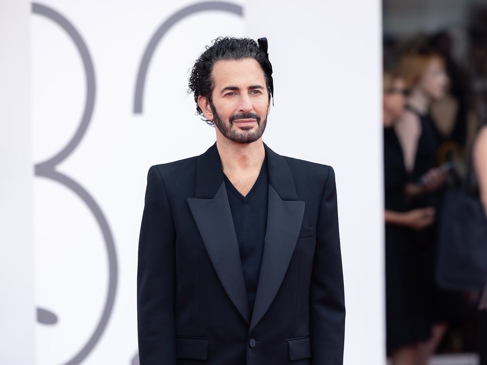 Marc Jacobs, bearded, his curly hair tied back with a black bow, in a black jacket with satin lapels and a black V-neck jumper, against a white background.