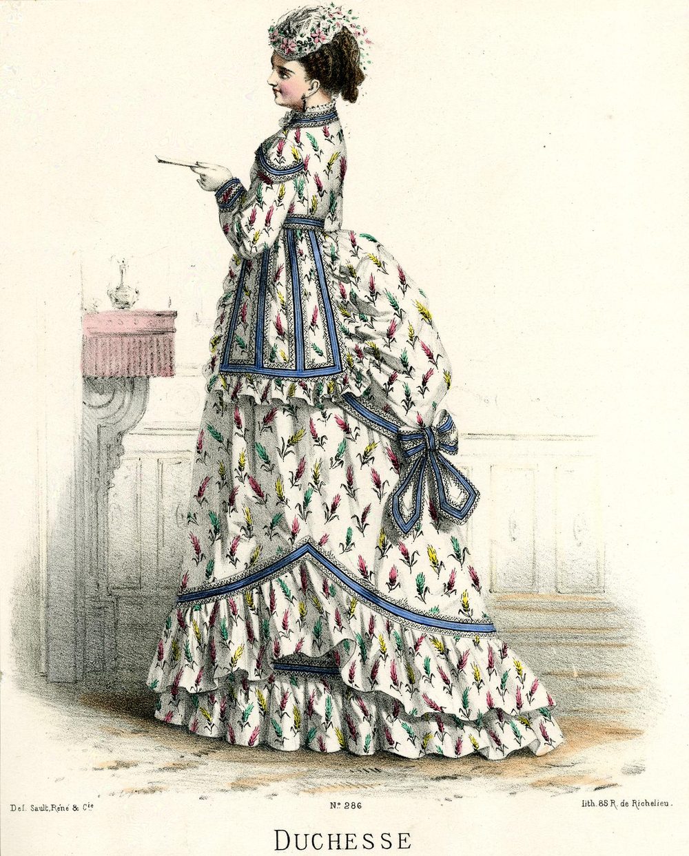 Hand-coloured fashion plate: a woman in profile in a white dress with colourful motifs and blue trim; a panel springs from the waist and falls over the skirt, with a bustle and a large bow at the back.