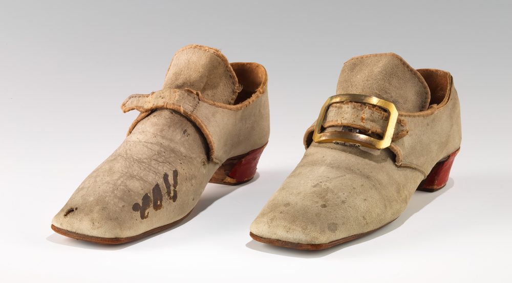 A pair of pale suede shoes with square toes, a gold buckle and red heels.