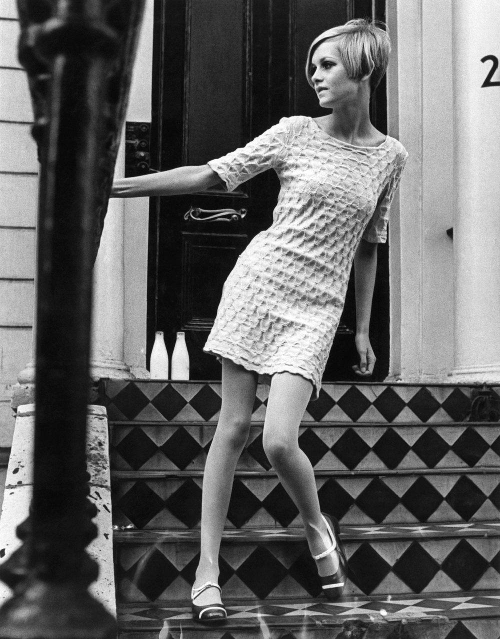 Black-and-white photo of a young woman with short blonde hair and a pale short-sleeved minidress, posing on chequered tiled steps in front of a black door, with two milk bottles by the threshold.