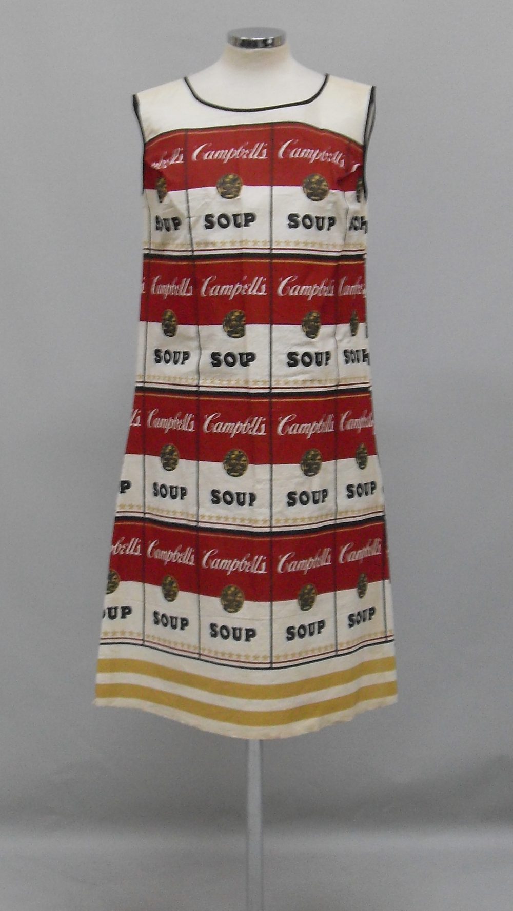 A sleeveless shift dress printed with rows of red-and-white Campbell’s soup cans and two gold stripes at the hem, on a dress form.