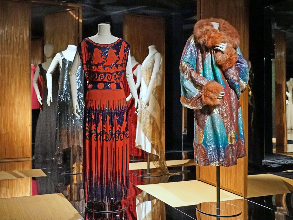 Garments on mannequins in an exhibition room with wooden panels: in the centre, a red dress embroidered with horses and waves in blue; on the right, an iridescent coat with a fur collar and cuffs; behind, a silver sequinned dress and a white one.