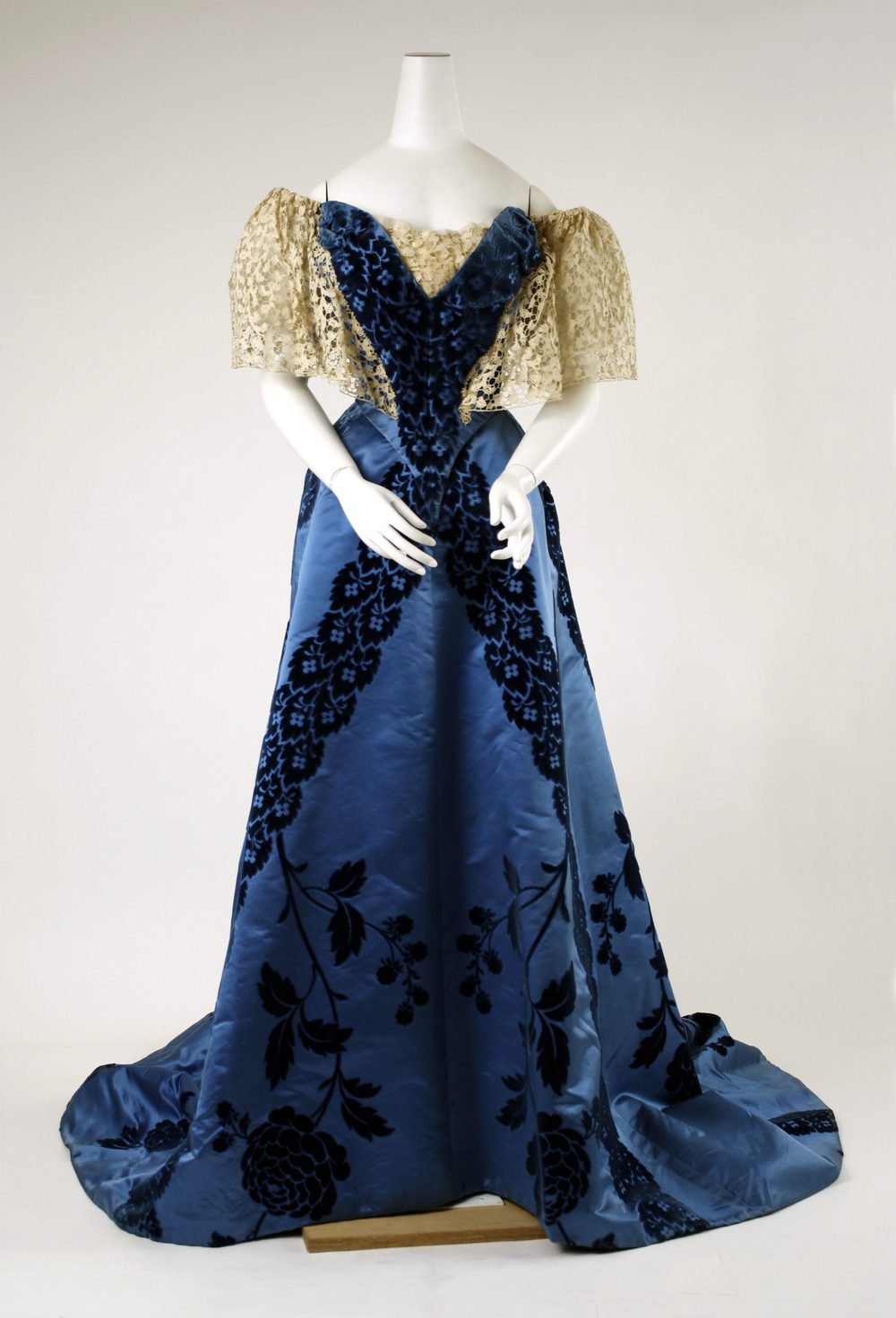 A blue satin evening dress with a train, dark blue floral garlands and short cream lace sleeves, on a white mannequin.