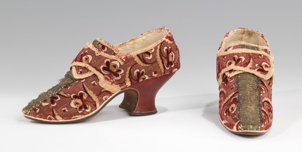 A pair of maroon fabric shoes with cream embroidered flowers, gold braid down the vamp and curved red heels; one seen in profile, the other from the front.